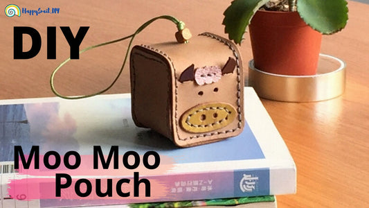 A box-shaped leather pouch with a cow pattern, attached to a keychain. The pouch is brown with a darker shade at the bottom, and there is a small plant beside it.