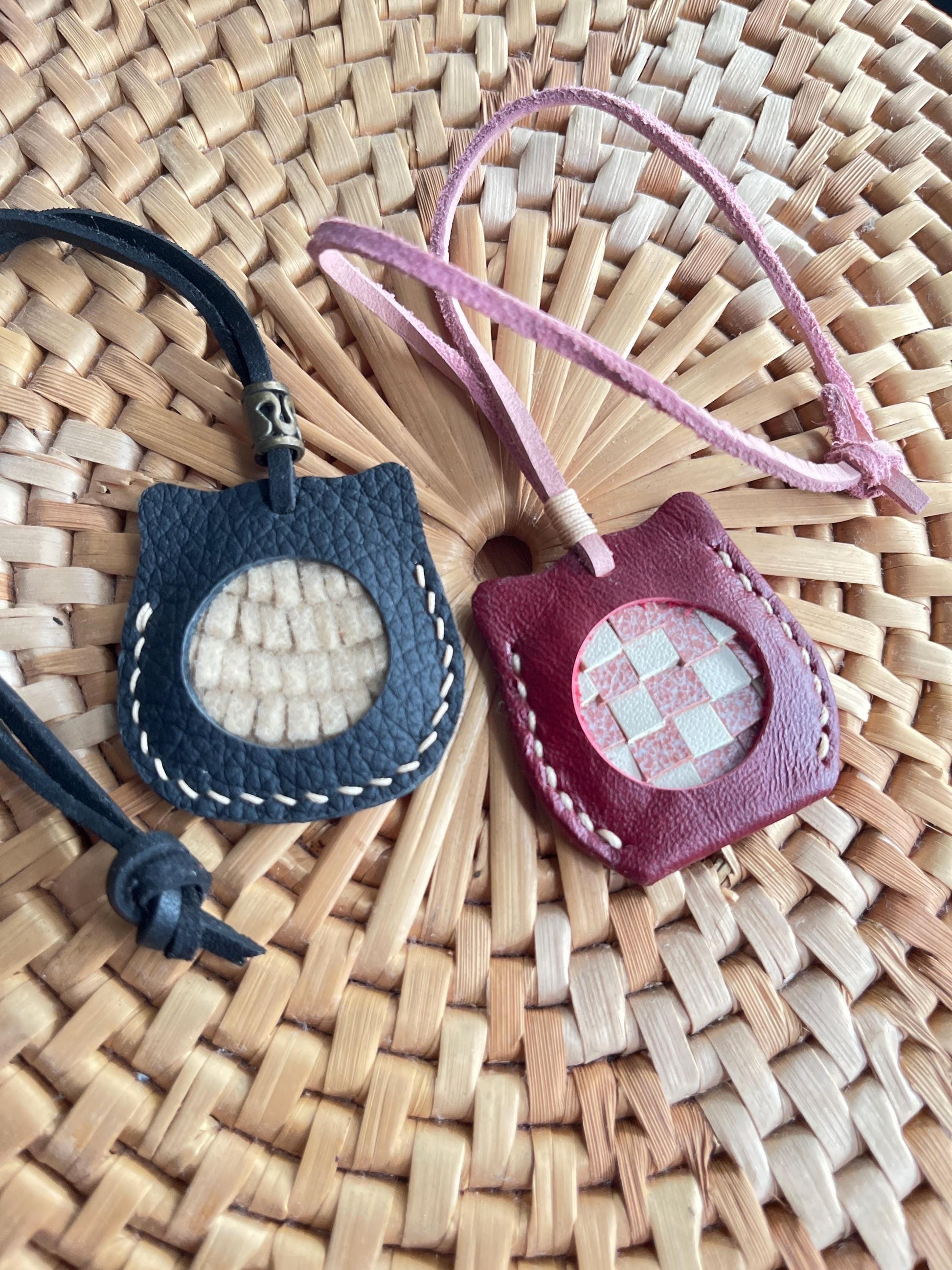 Two leather cat ornament  with woven fabric inserts. The leather cat ornament on the left is black with a beige insert and a silver clasp. The leather cat ornament on the right is burgundy with a white and red 