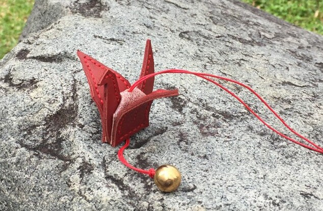Red leather origami crane with a gold bell charm attached to a red cord.