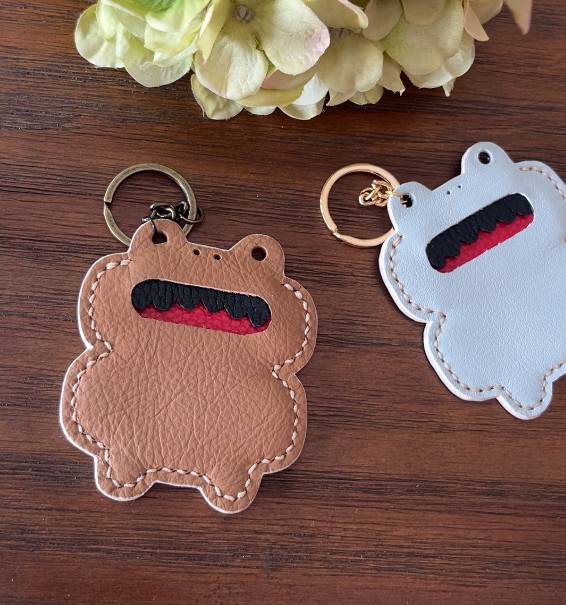 Two frog-shaped keyrings. One is brown leather with a black and red mouth, the other is light blue. Both have metal key rings and white stitching around the edges. The keyrings are on a wooden surface with a flower in the background.