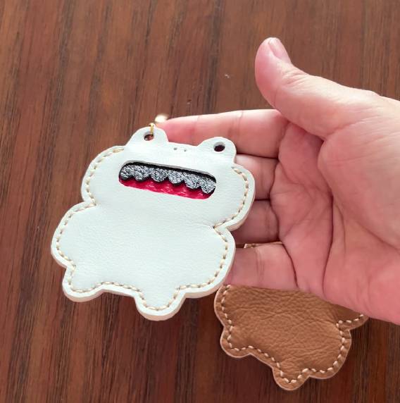A white leather frog-shaped keyring with a red and black mouth design. The keyring has a gold-coloured ring for attachment and is stitched with light brown thread. A second, brown leather piece is visible.