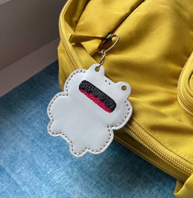 A white leather frog-shaped keyring with a red and black mouth design. It features a gold chain and a black and pink design. The keyring is attached to a yellow backpack.