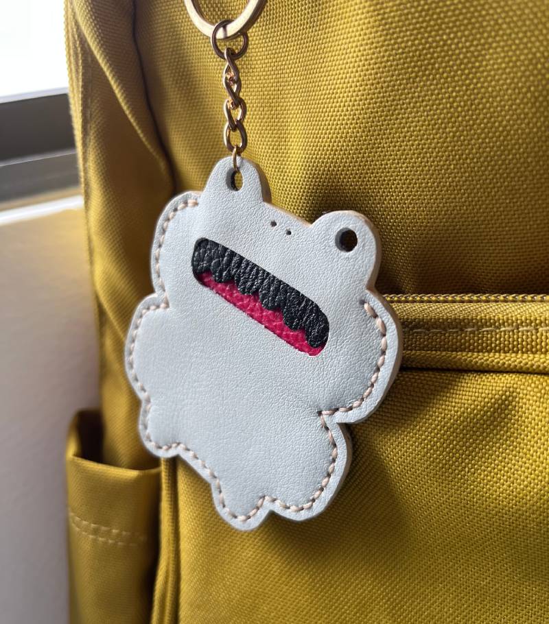 Another view of A white frog-shaped keyring with a red and black mouth hangs from a gold-colored chain attached to a yellow backpack. The backpack has multiple pockets and a zip closure.
