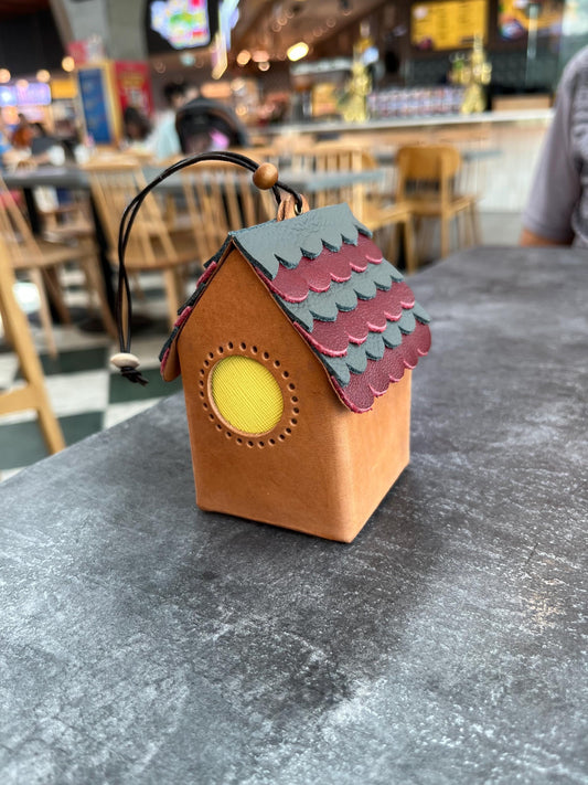 A mini brown leather house-shaped ornament with a red and black roof. The roof has a scalloped design and the house has a yellow circle window.
