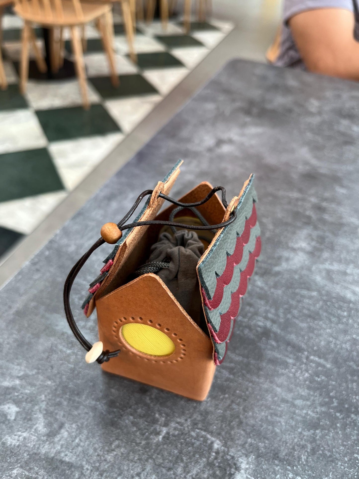 A mini, brown leather pouch shaped like a house with a red and blue patterned roof. The pouch has a yellow circle window and a drawstring closure.
