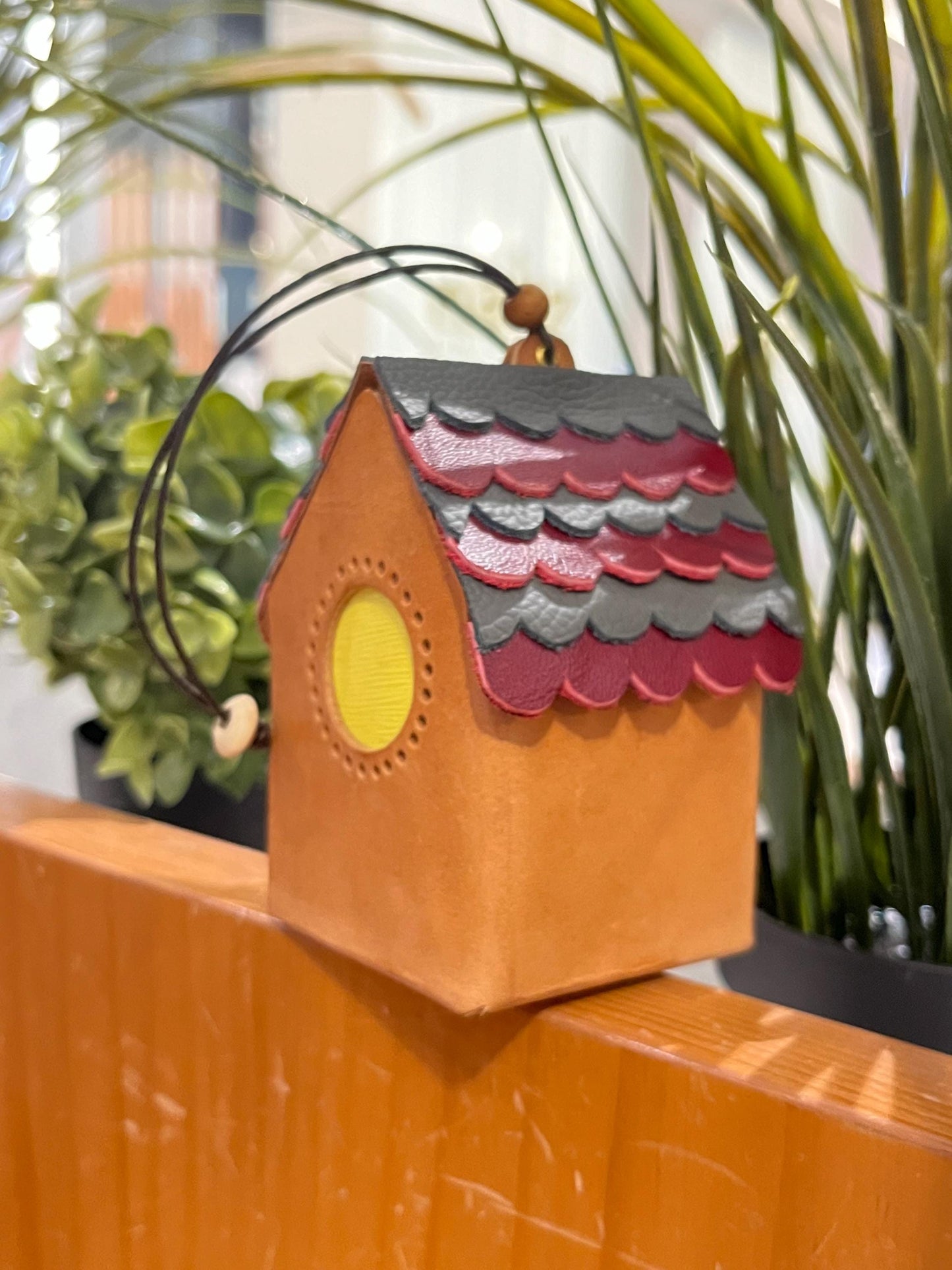 A mini, brown leather house-shaped ornament with a red and black roof. The roof is made of overlapping scales and has a yellow circle window.