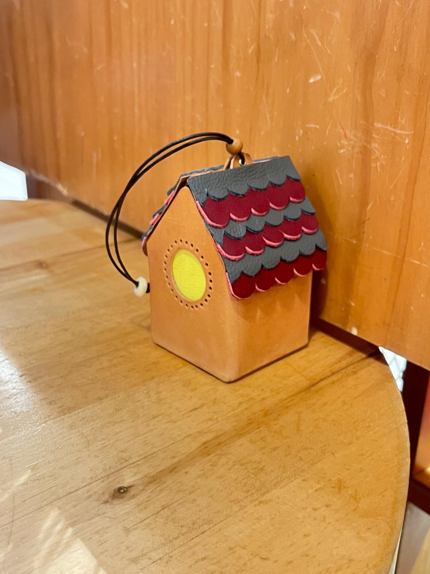 A mini, brown leather house-shaped keyring with a red and grey roof. The roof has a scalloped design and a yellow circle window.