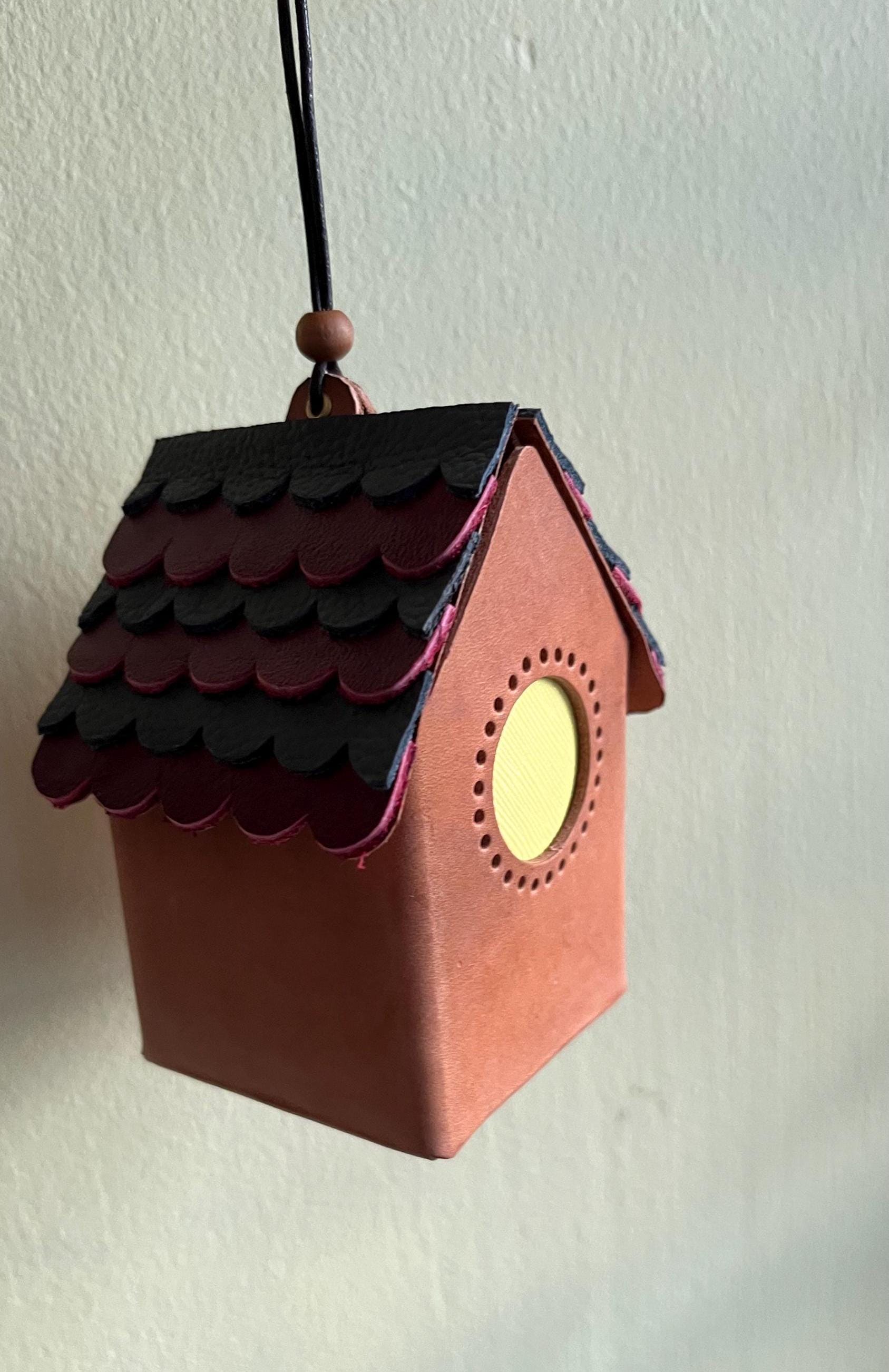 A mini, red leather house-shaped ornament with a black and burgundy roof and a yellow window. The ornament is hanging from a black cord.