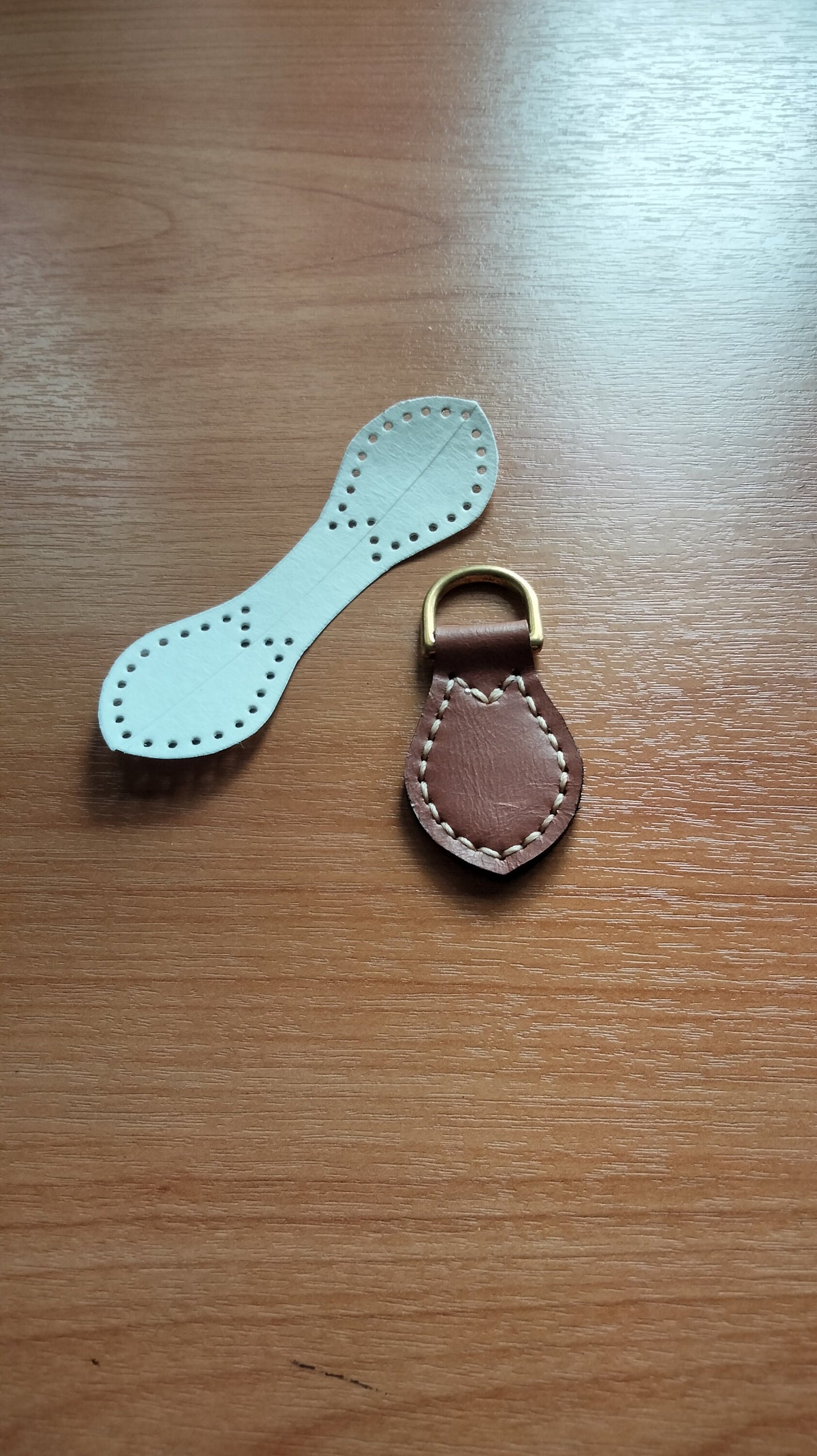 A white leather strap with holes for stitching and a brown leather loop with a gold metal ring and its leather pattern on the table.