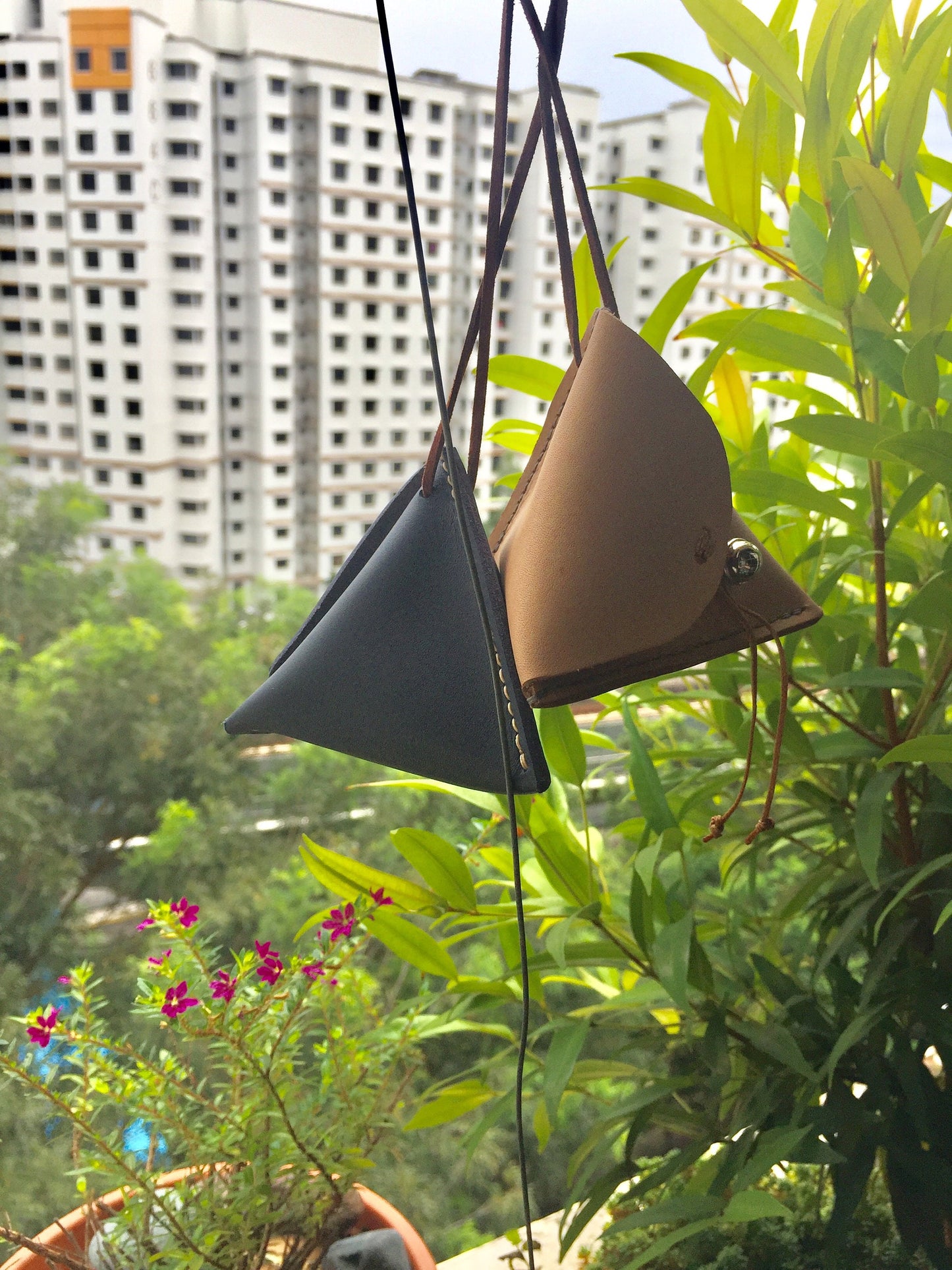 Two small pyramid Leather coin pouches hanging from a string. The pouches are triangular in shape and are made of dark blue and brown leather. The pouches are hanging in front of a window with a view of a building.