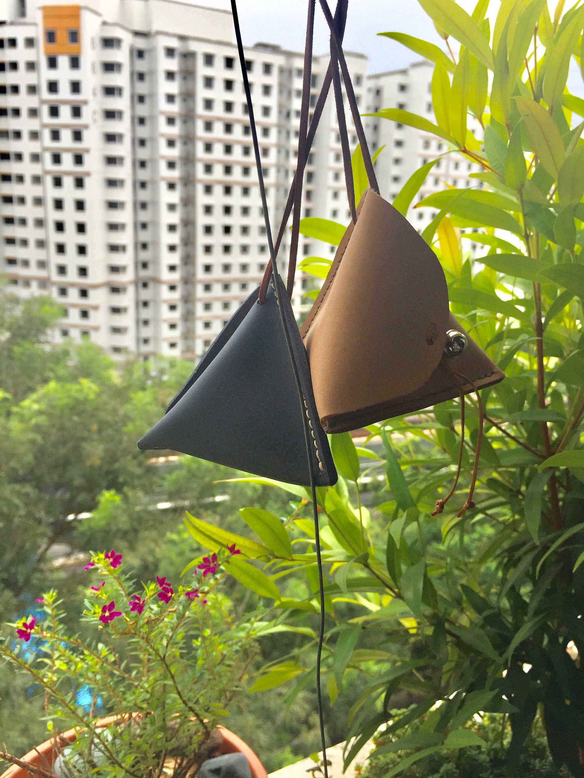 Two small pyramid Leather coin pouches hanging from a string. The pouches are triangular in shape and are made of dark blue and brown leather. The pouches are hanging in front of a window with a view of a building.