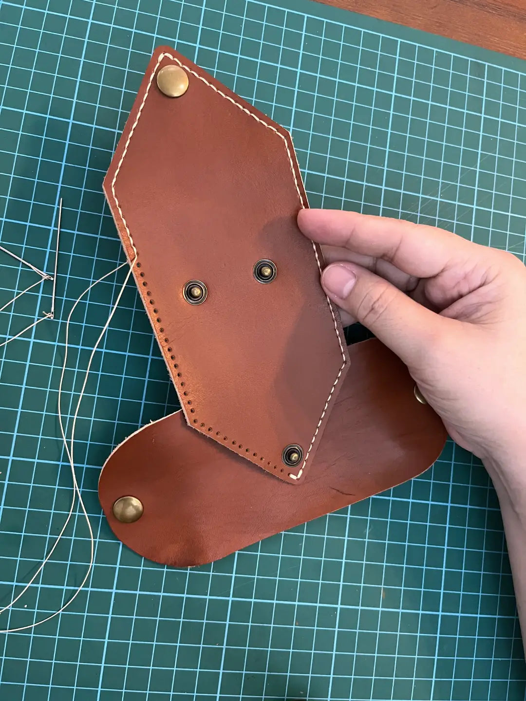 Stitching a brown leather coin pouch with gold studs on a green cutting mat