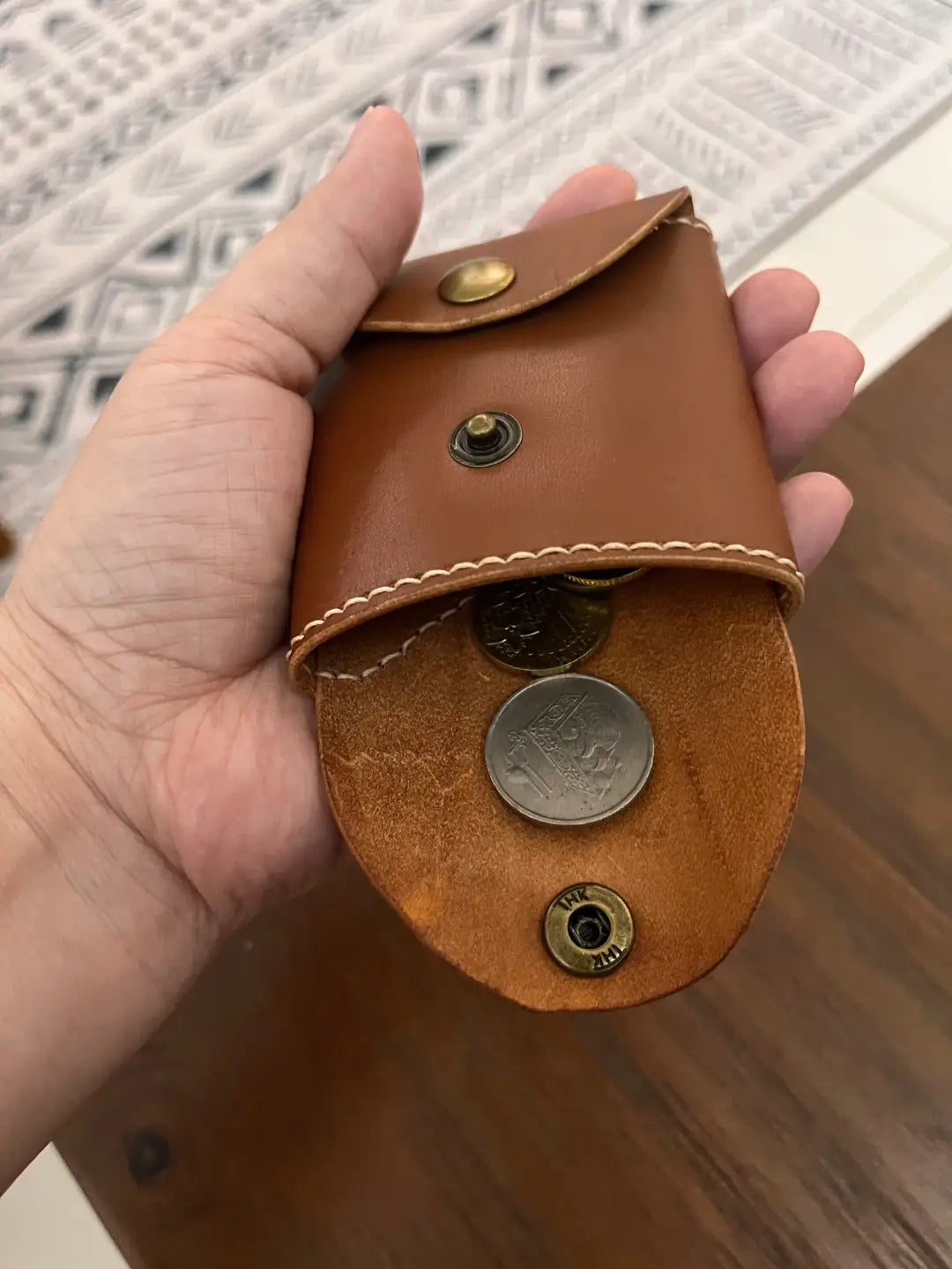 Hand holding a brown leather envelope shape coin pouch with coins inside against a patterned surface.