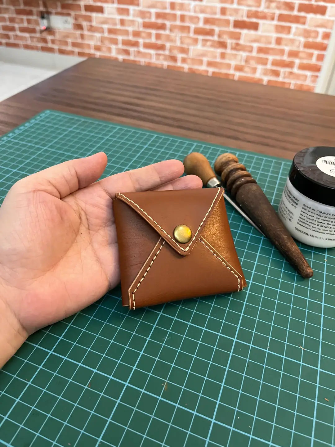 Hand holding a small brown leather  coin wallet with a yellow button on a green cutting mat.