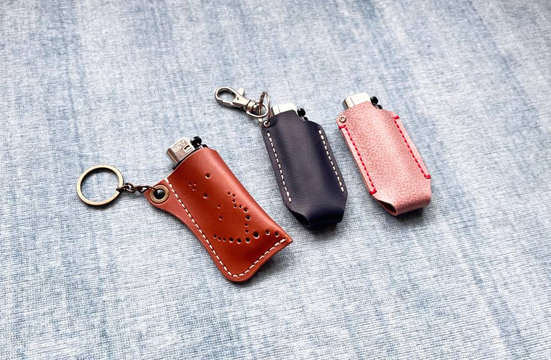 Three slim leather cases with keychain in brown, black, and pink on a textured gray surface.