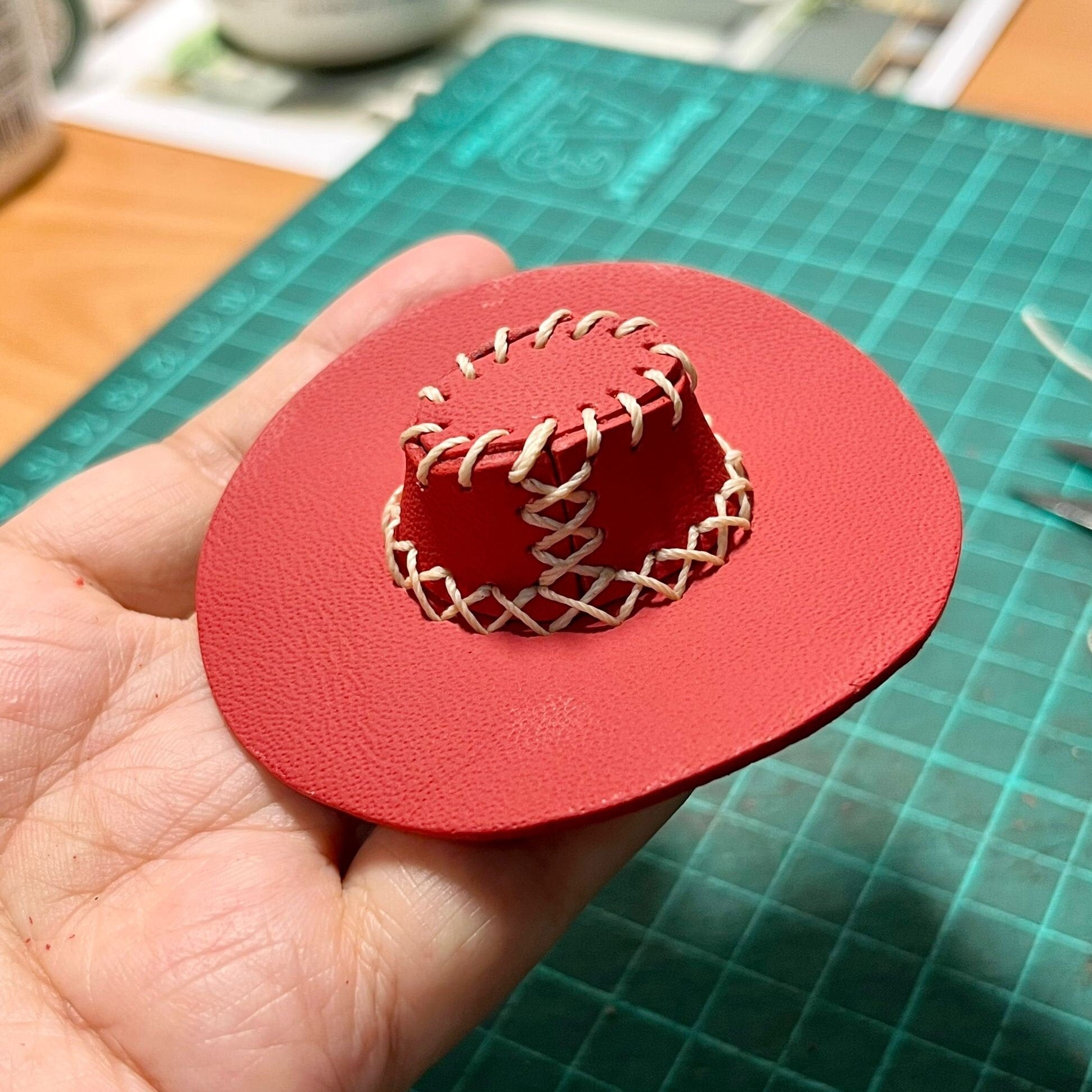 Unique leather gift for western lovers featuring a tiny, realistic cowboy hat design. Hand-sewn artisan leather miniature cowboy hat with professional stitching. A small, red leather hat with a wide brim and a cylindrical crown. The crown is stitched with cream-coloured thread in a decorative pattern. The hat is a handcrafted item, possibly for a doll or small figurine.